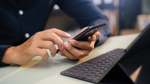 A young man surfing on a phone sitting in front of a tablet. 