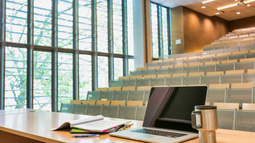 Photo of laptop on a professor's desk with tumbler in the front of a lecture hall